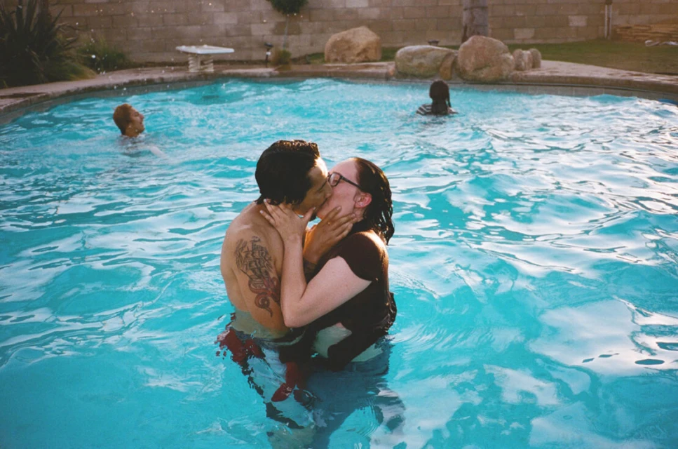 A couple kisses in a blue outdoor swimming pool with other people in the background, set against a concrete block wall.