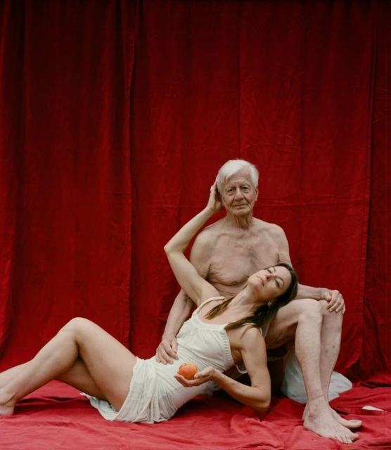 Elderly man and woman in a red-toned studio portrait with an apple. The female model reclines against the seated older man.