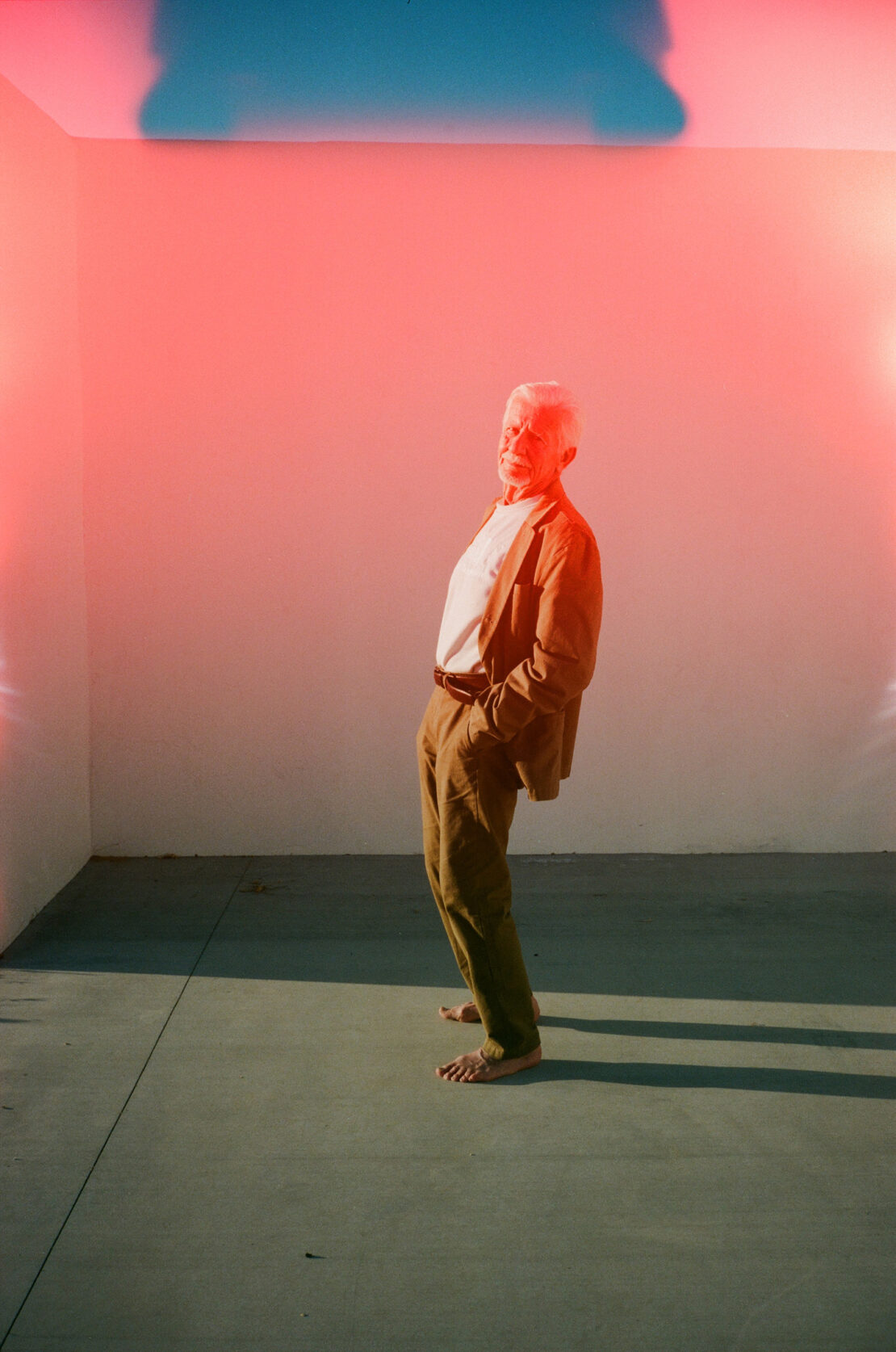 Elderly man in a suit standing barefoot in a colorfully lit empty room, posing with his hands in his pockets against a white wall.