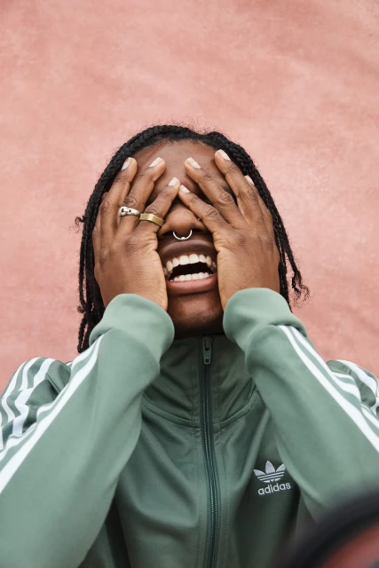 Young adult covering face, mouth open, wearing Adidas tracksuit and rings against textured pink wall in a medium shot, studio lighting.