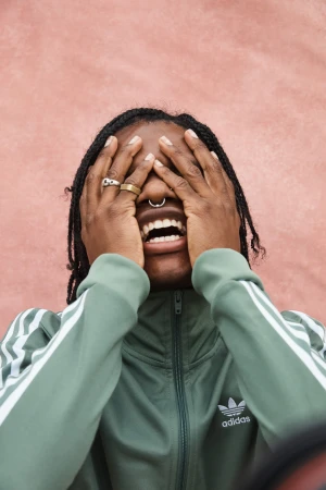 Young adult covering face, mouth open, wearing Adidas tracksuit and rings against textured pink wall in a medium shot, studio lighting.