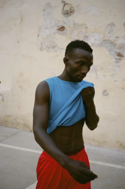 Portrait of a Black man in a blue tank top and red shorts, looking down against a tan wall with peeling paint in natural light.