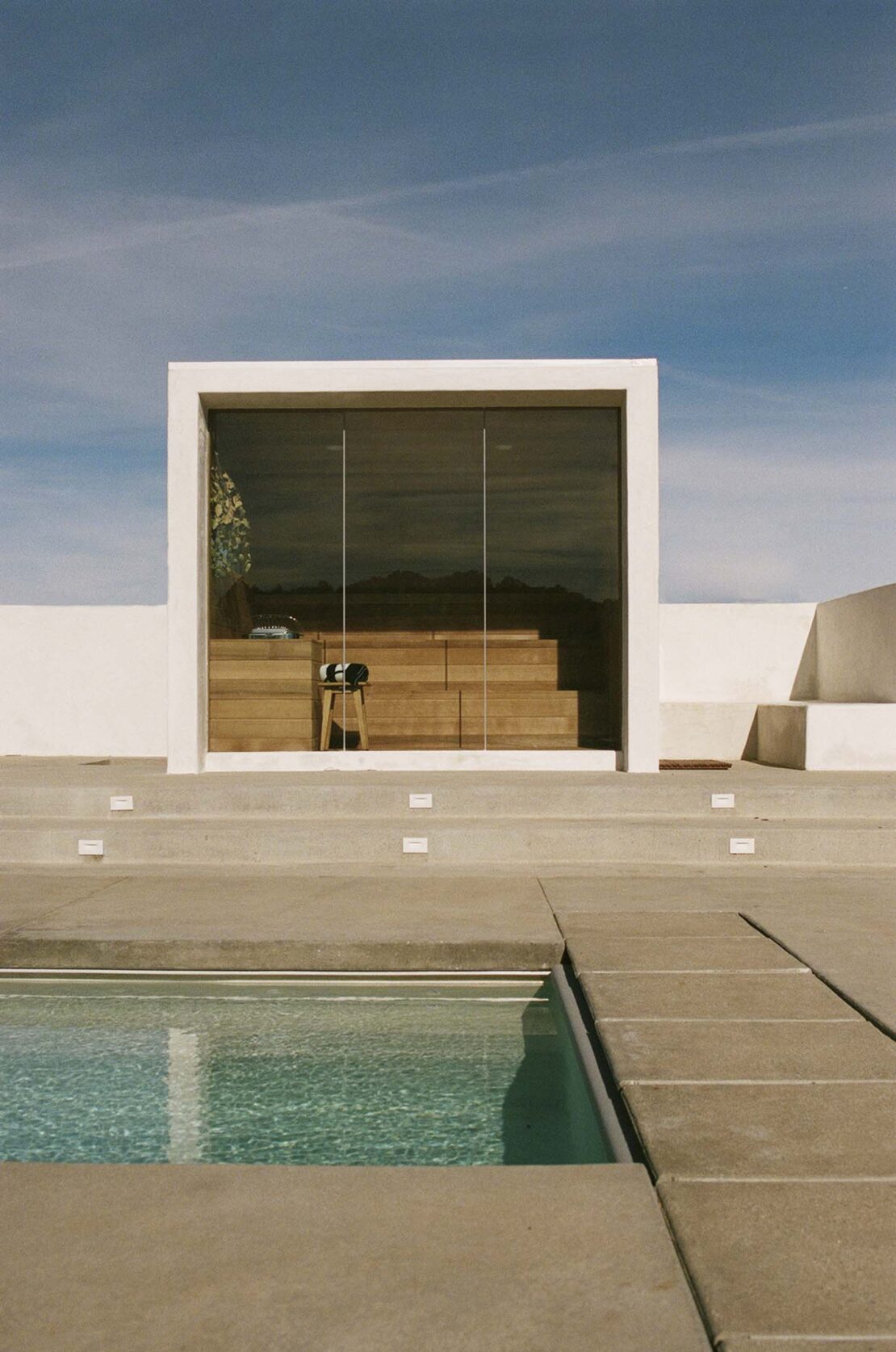 Exterior view of a modern minimalist white building with a glass-enclosed living space, concrete steps, and a swimming pool on a sunny day.