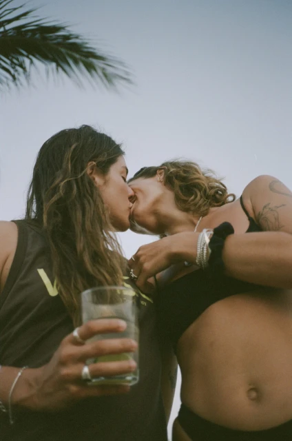 A loving lesbian couple shares a kiss at an outdoor celebration, set against a vibrant sky, with a clear drink and palm fronds.