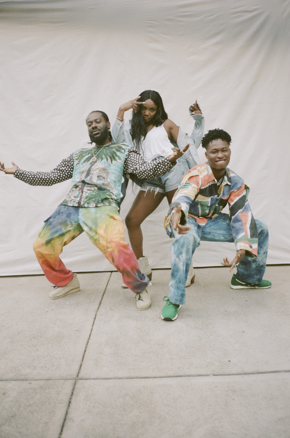 Three people strike poses indoors against a bright backdrop. The trio sports vibrant, casual ensembles with patterned tops and colorful pants.