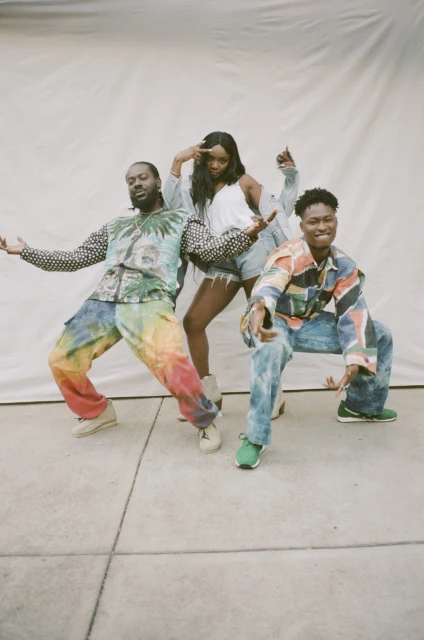 Three people strike poses indoors against a bright backdrop. The trio sports vibrant, casual ensembles with patterned tops and colorful pants.