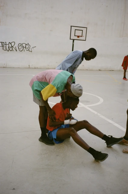 Three young Black men in an indoor basketball court, one sitting with a bandaged wrist, another attending to him, a basketball hoop in the background.