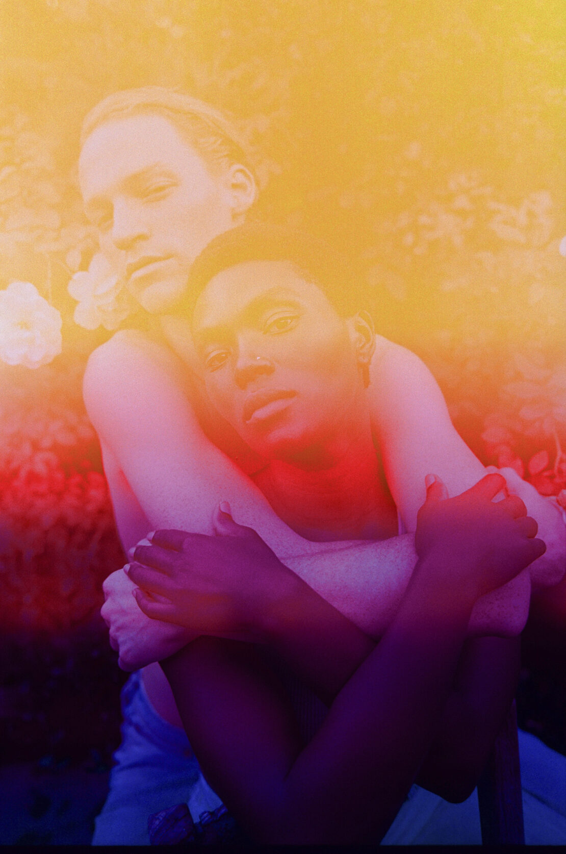 A couple embraces outdoors with artistic lighting: one person embraces the other from behind, both looking at the camera.