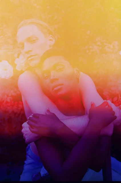 A couple embraces outdoors with artistic lighting: one person embraces the other from behind, both looking at the camera.