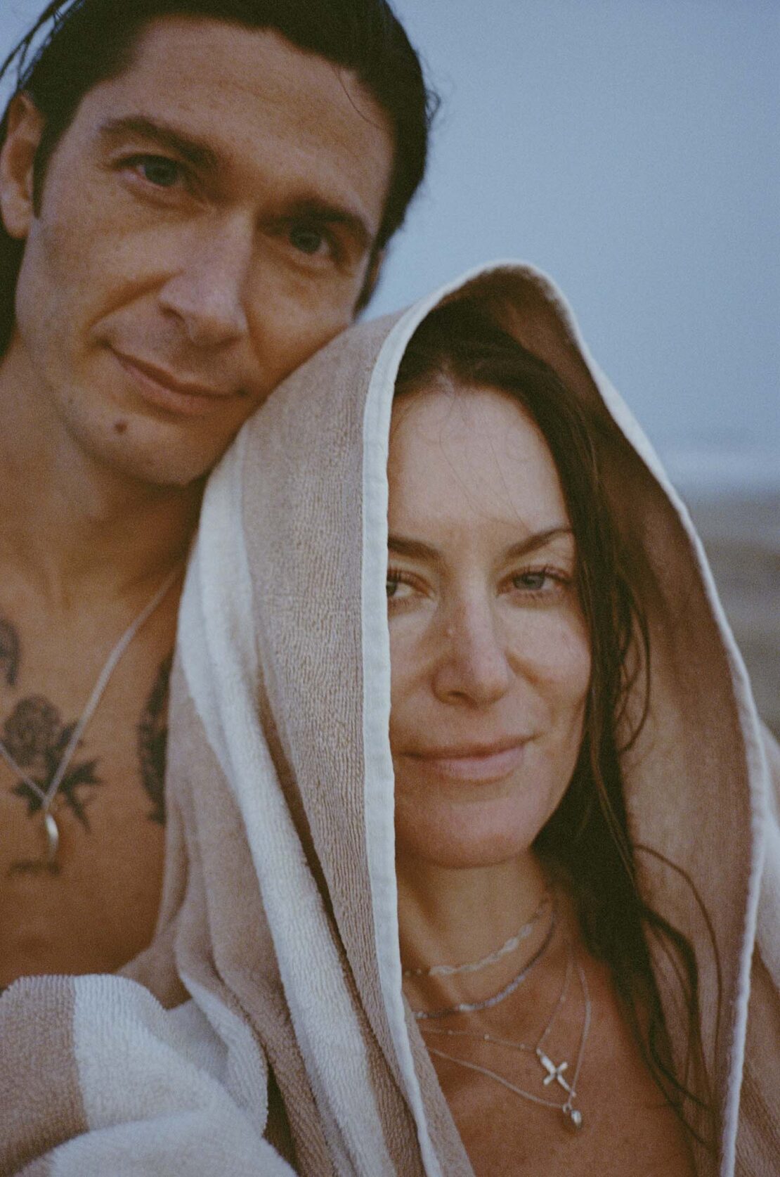 A couple poses closely on a beach, the woman's head covered by a striped towel, under a blue sky. The man has visible tattoos.