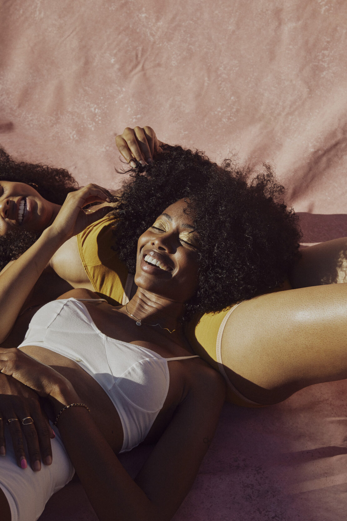 Two smiling Black women in lingerie lie close together on a pink background, showcasing natural hair, glowing skin, and relaxed joy.
