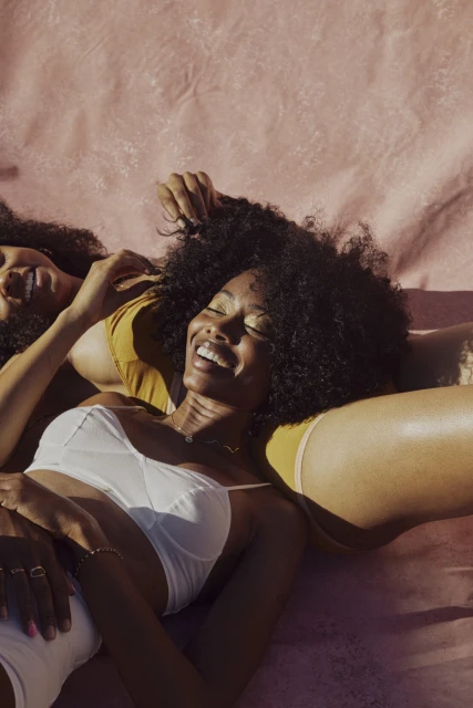 Two smiling Black women in lingerie lie close together on a pink background, showcasing natural hair, glowing skin, and relaxed joy.
