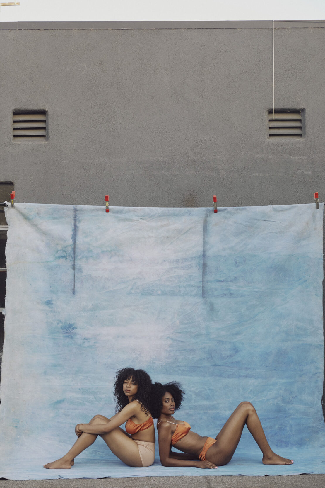 Two Black women pose back-to-back in orange bikinis against a blue cloth backdrop outdoors. The setting is urban with a grey wall.