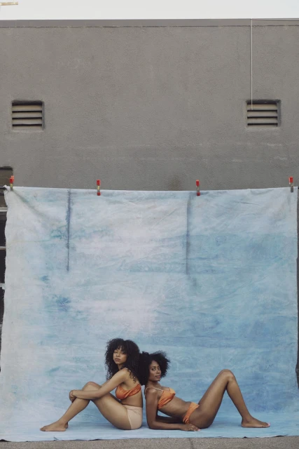 Two Black women pose back-to-back in orange bikinis against a blue cloth backdrop outdoors. The setting is urban with a grey wall.