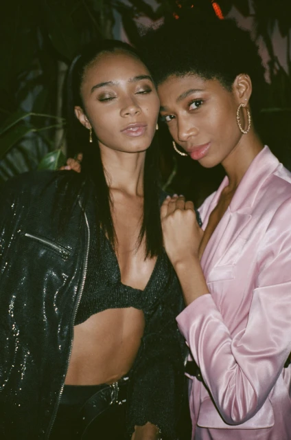 Two Black women pose indoors in fashionable attire, one in a pink jacket and the other in a black sequin jacket and bra top.