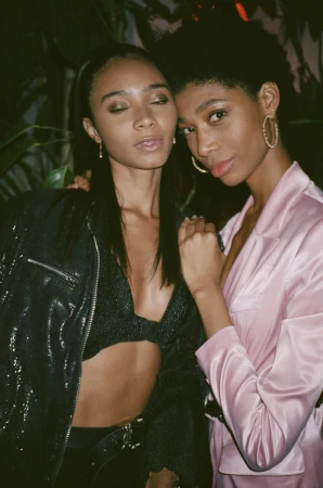 Two Black women pose indoors in fashionable attire, one in a pink jacket and the other in a black sequin jacket and bra top.