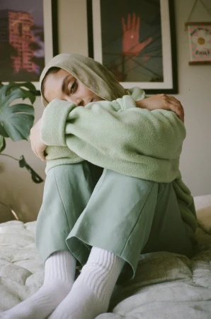 Woman in sage green hoodie and trousers hugging her knees on a bed by Magdalena Wosinska