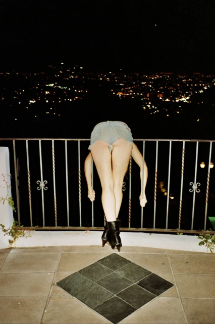 A woman with long brown hair and a black jacket poses in front of a blurry, brightly lit background for a fashion shot.
