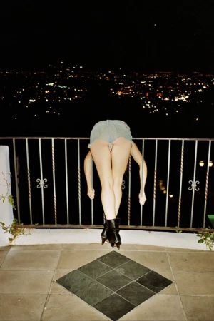 A woman with long brown hair and a black jacket poses in front of a blurry, brightly lit background for a fashion shot.