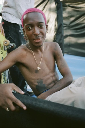 Young man with short pink-dyed hair and a gold chain necklace seated outdoors, captured in a candid portrait style.