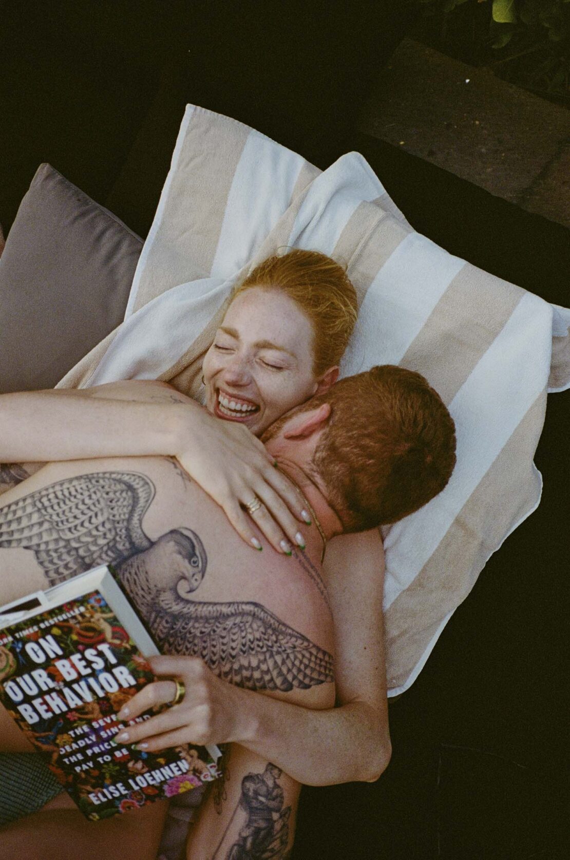 Woman with red hair smiles while being embraced by a man with tattoos; book titled 'On Our Best Behavior' is visible in the foreground.