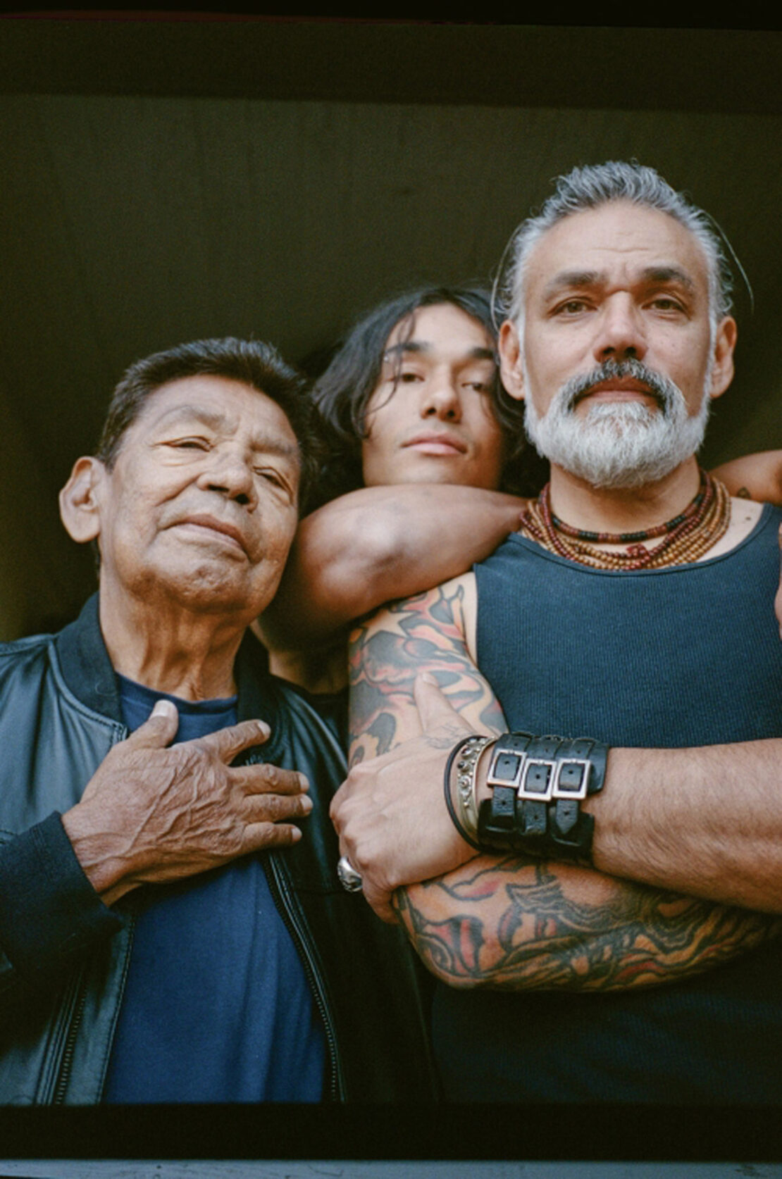 Portrait of a three-generation family of Indigenous men standing together indoors with natural light on their faces and shoulders.