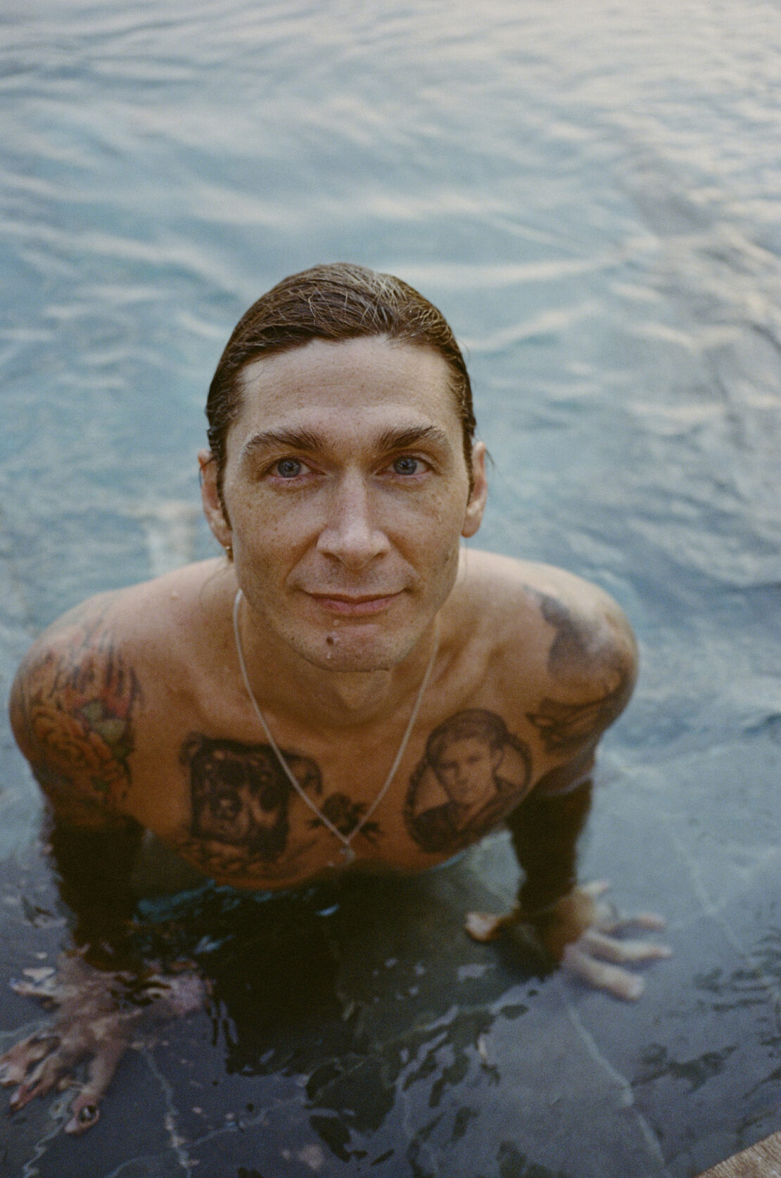 Man with tattoos on chest and arms submerged in swimming water looking up at the camera by Magdalena Wosinska