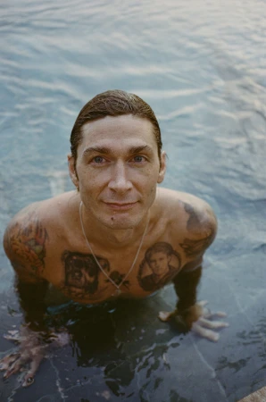 Man with tattoos on chest and arms submerged in swimming water looking up at the camera by Magdalena Wosinska