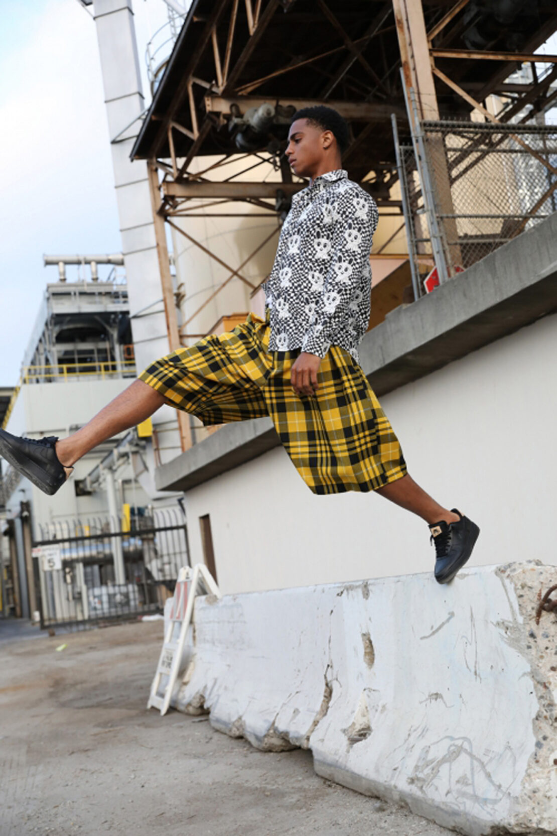An adult male in yellow plaid shorts and a skull-patterned shirt leaps over a concrete barrier outdoors in an urban industrial environment.