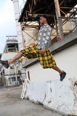 An adult male in yellow plaid shorts and a skull-patterned shirt leaps over a concrete barrier outdoors in an urban industrial environment.