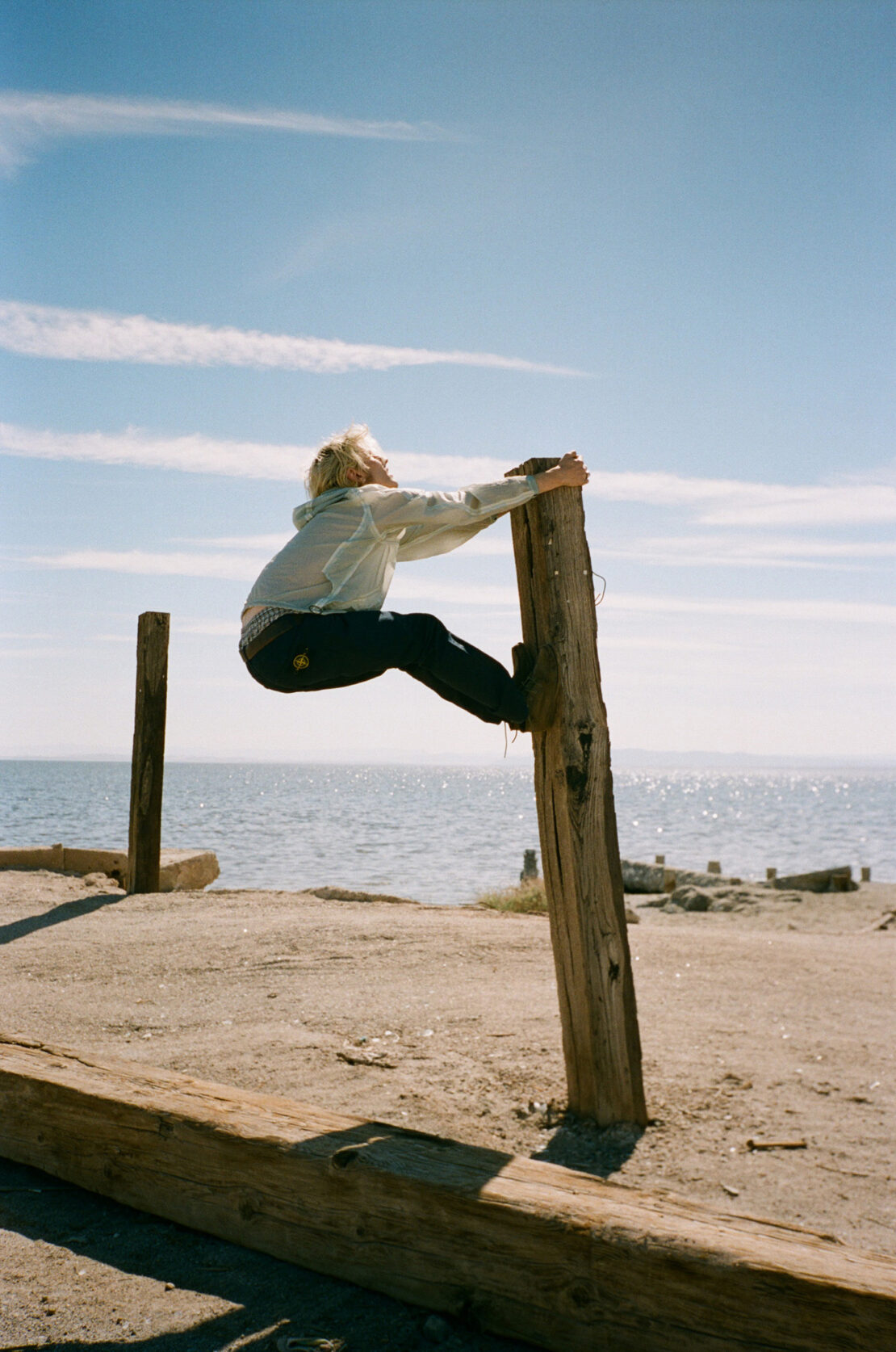 A person in a light jacket balances on a wooden post with the ocean and sky in the background on a sunny day in an outdoor setting.