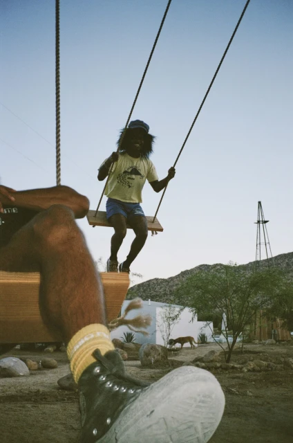 Man on a wooden swing in a desert landscape with a second person partially visible. A building and distant mountains are visible.