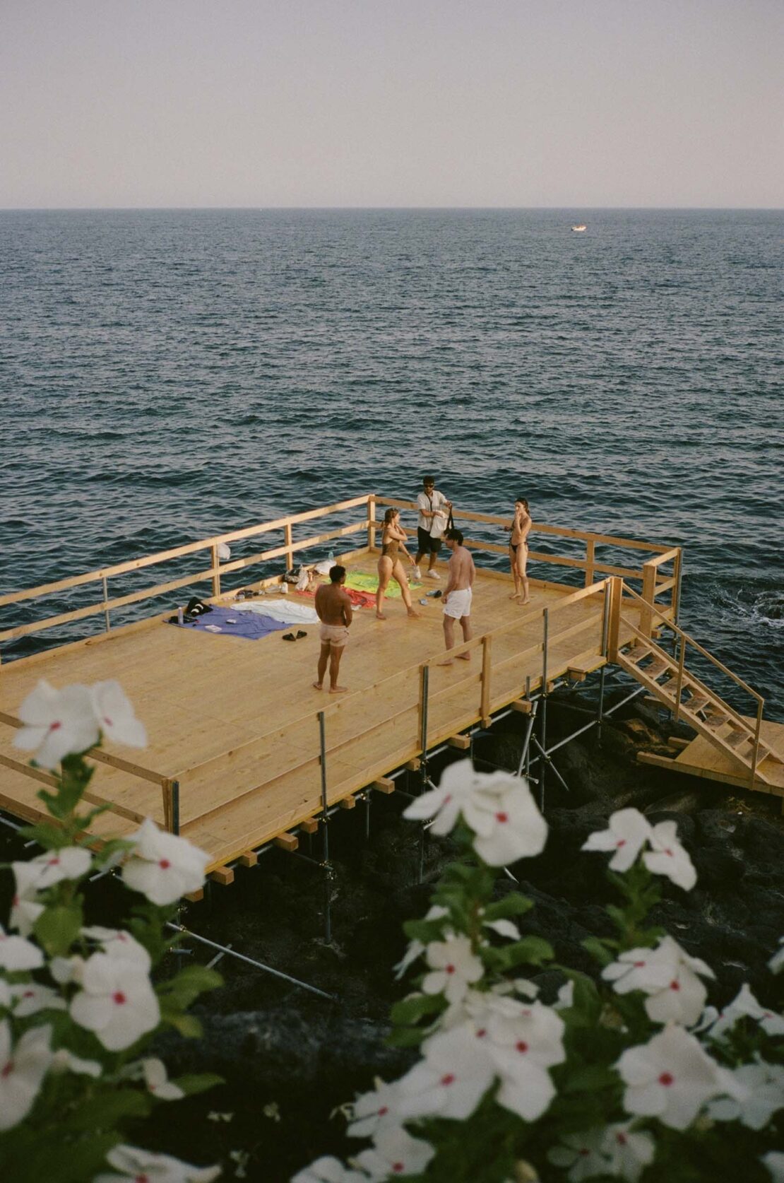 Group of people on a wooden deck by the ocean. Some are wearing swimwear; the sky is clear, and flowers are visible in the foreground.
