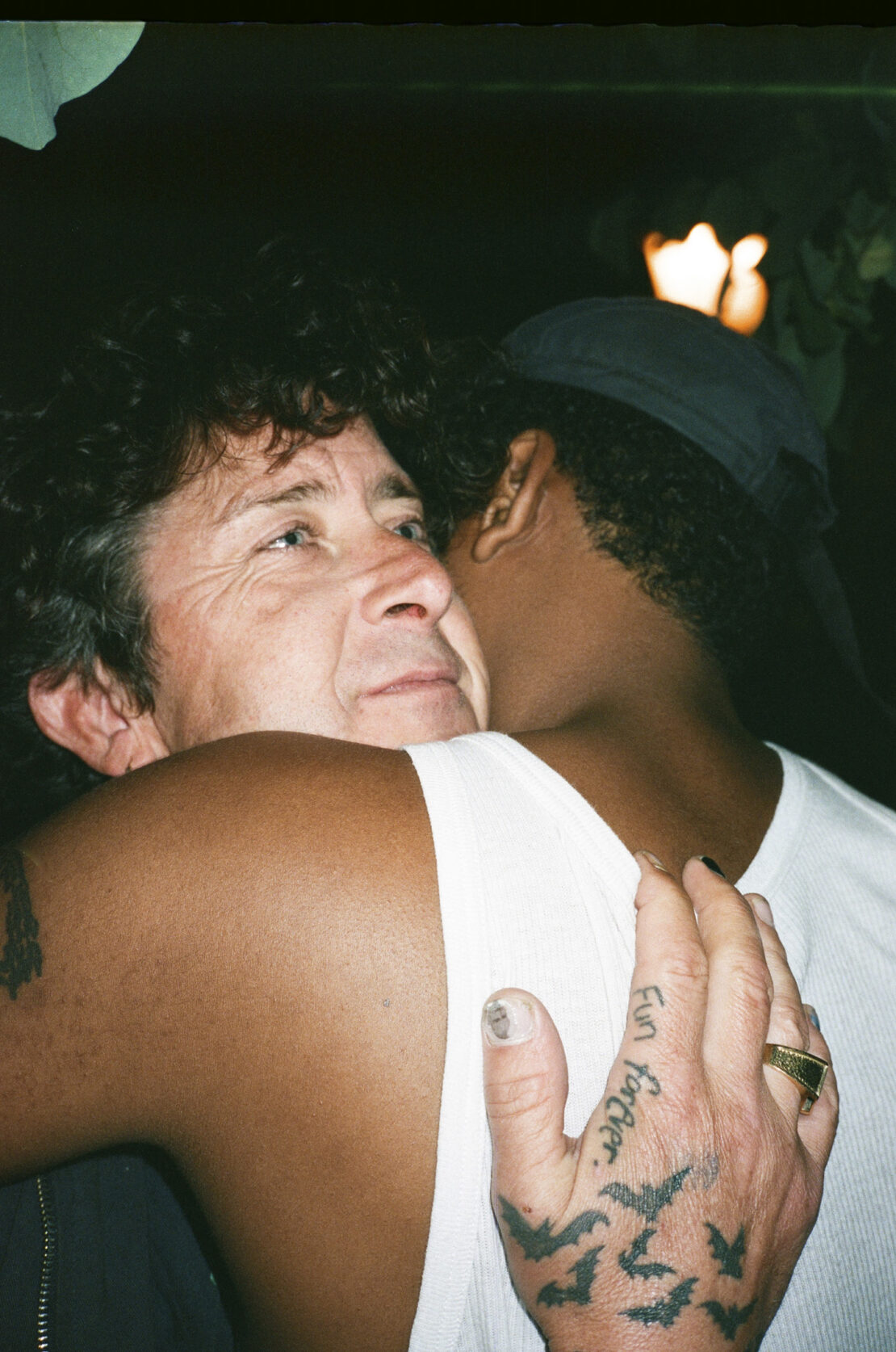Close-up of two people embracing, one with dark hair and cap, the other with curly hair and tattoos, indoor setting with dark lighting.