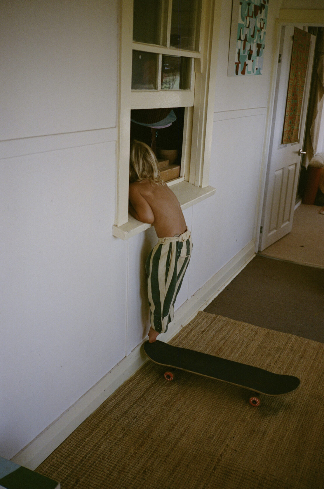 Child peers through window in striped pants standing on skateboard inside. Interior shot with natural light, capturing a candid moment.