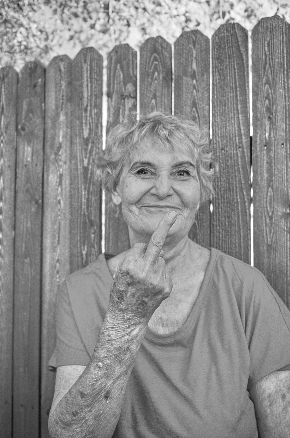 Monochrome portrait of an elderly woman smiling with curly hair, holding up her middle finger in front of a wooden fence.