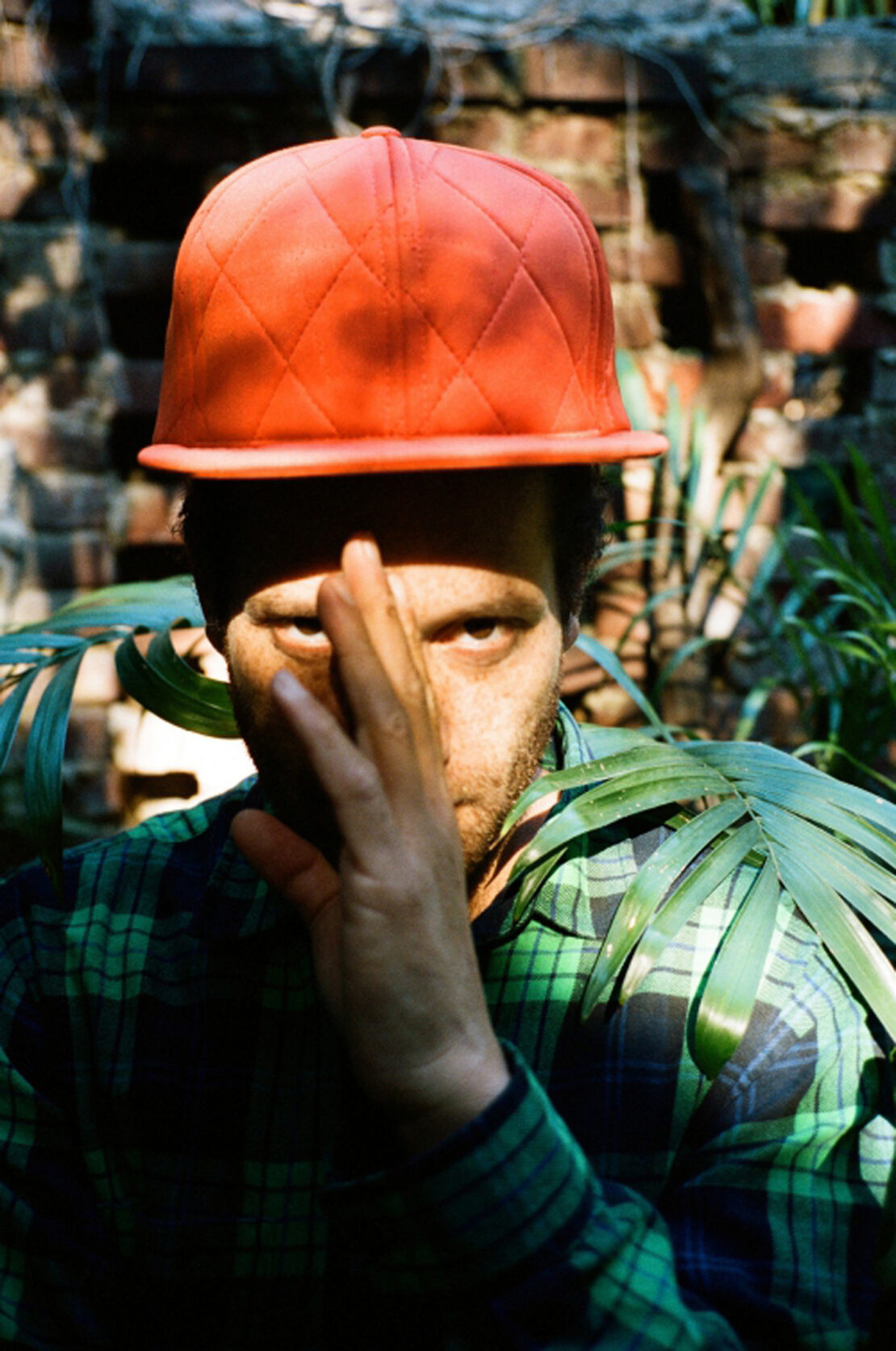 An adult with stubble wearing an orange baseball cap poses in front of a brick wall with foliage. The subject partially obscures his face.