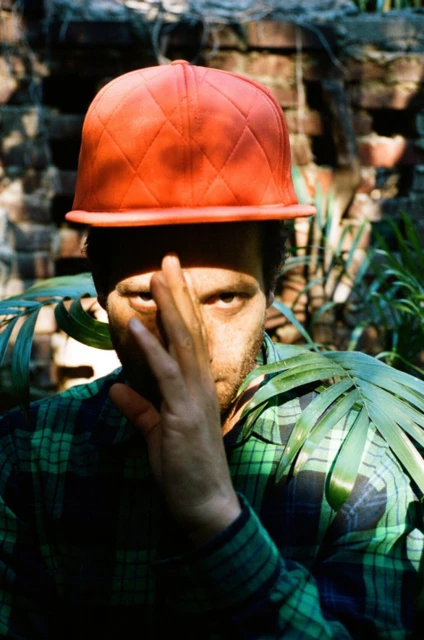 An adult with stubble wearing an orange baseball cap poses in front of a brick wall with foliage. The subject partially obscures his face.