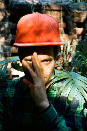 An adult with stubble wearing an orange baseball cap poses in front of a brick wall with foliage. The subject partially obscures his face.