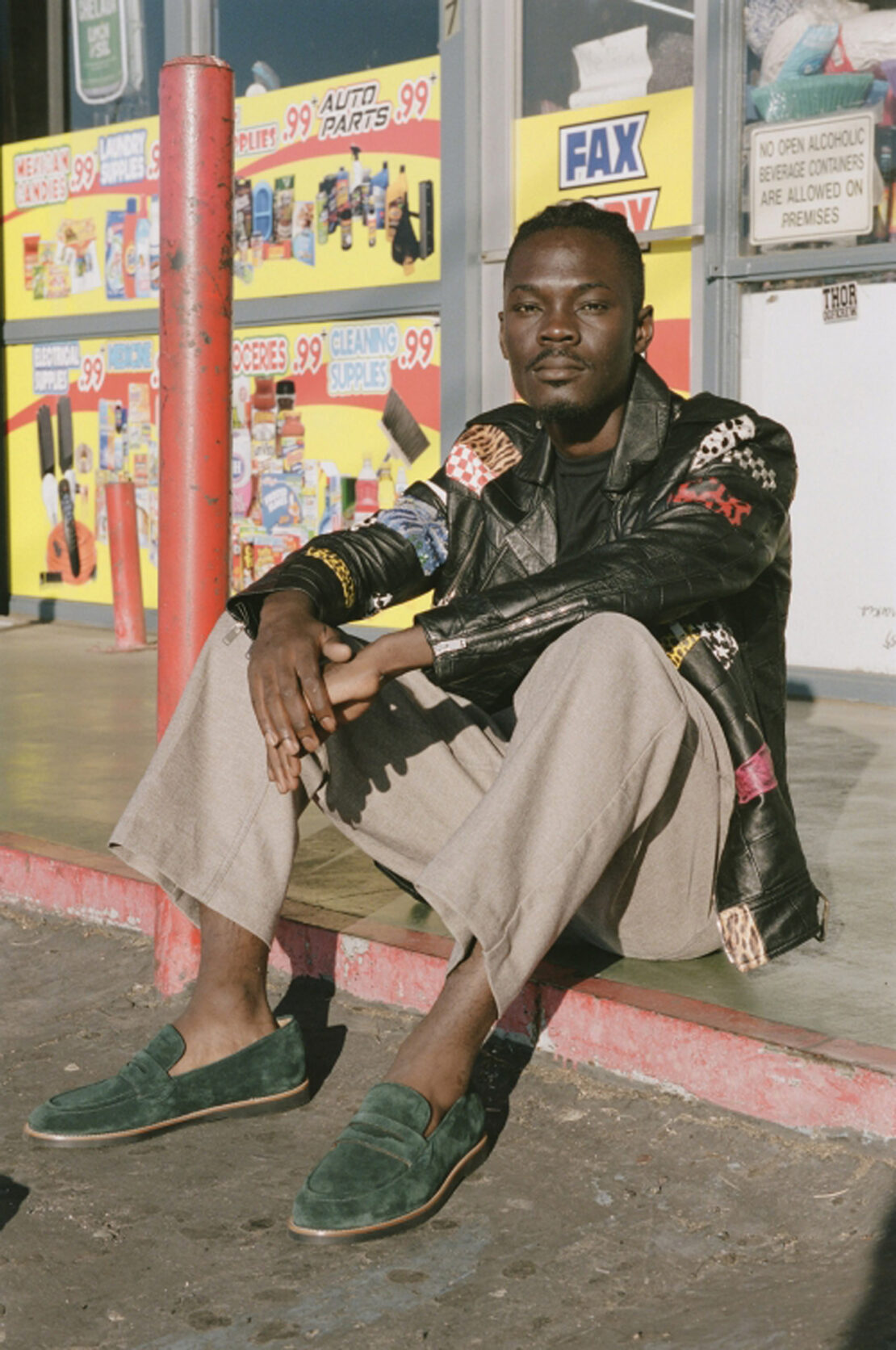 Black man wearing a patterned leather jacket and green suede loafers seated outdoors by a storefront