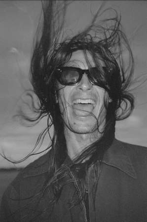 Portrait of a laughing person wearing dark sunglasses with long dark hair blowing wildly in the wind by Magdalena Wosinska