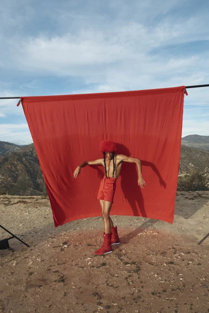 Adult male model in a red outfit poses with arms outstretched against a red backdrop, set on a desert landscape under a bright blue sky.