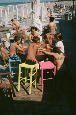 A group of children sit around a round table on a deck at the beach, with lounge chairs and the ocean visible in the background.