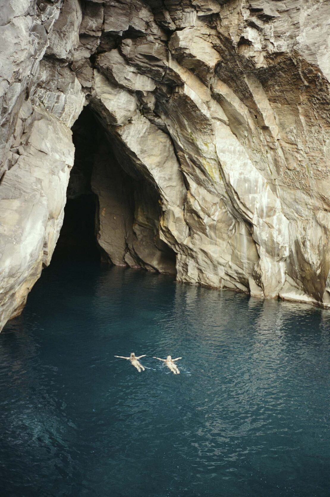 Two people float on their backs in bright blue water inside a large cave opening with light reflecting on the rock walls.