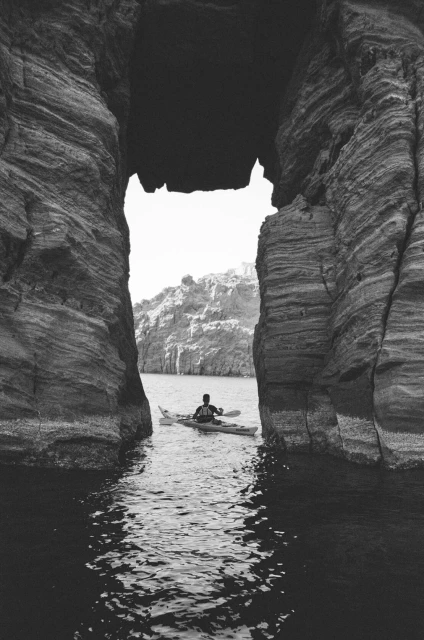 A person in a kayak paddles through a natural rock archway in black and white. Water ripples lead the eye to the paddler.