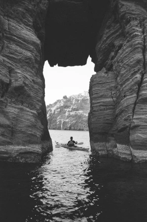 A person in a kayak paddles through a natural rock archway in black and white. Water ripples lead the eye to the paddler.