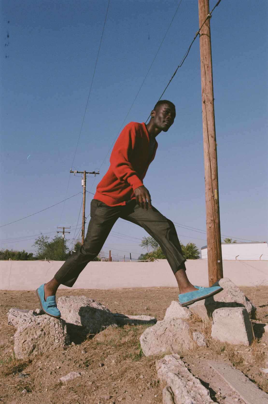 Young man in red sweater and turquoise shoes strikes pose on concrete rubble under blue sky with utility pole in background.