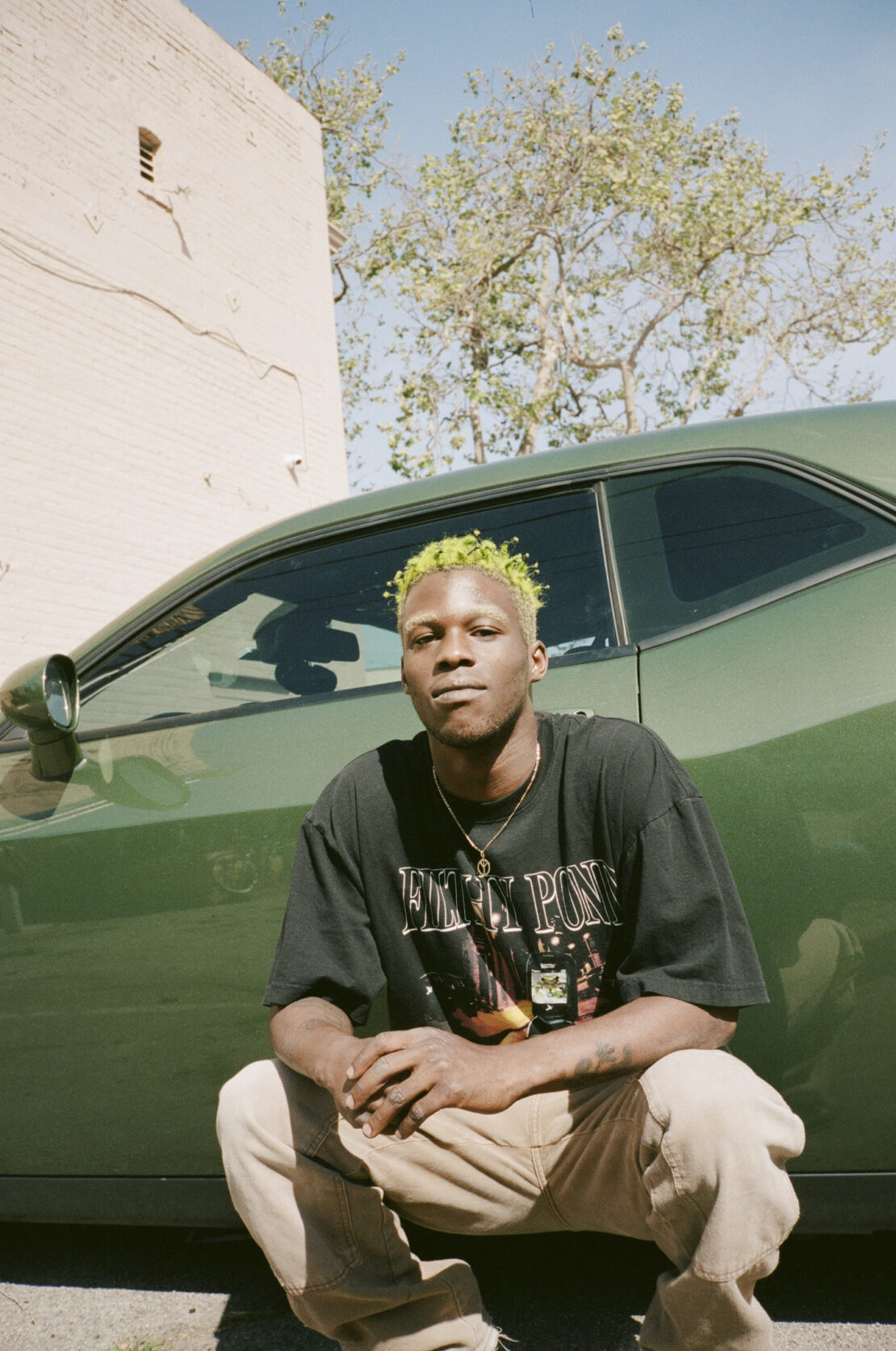 Portrait of a young man with bleached hair crouching in front of a green car, wearing a graphic t-shirt and tan pants.