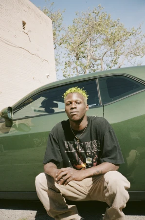 Portrait of a young man with bleached hair crouching in front of a green car, wearing a graphic t-shirt and tan pants.
