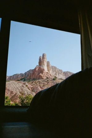 A rocky mountain landscape framed through a window, with a curtain visible on the right side and a clear blue sky above.
